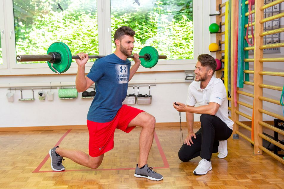 Individuelles und zielgerichtetes Personaltraining. Patient mit einer Langhantel auf der Schulter macht eine Ausfallschrittbewegung nach vorne, der Physiotherapeut betrachtet die Ausführung.