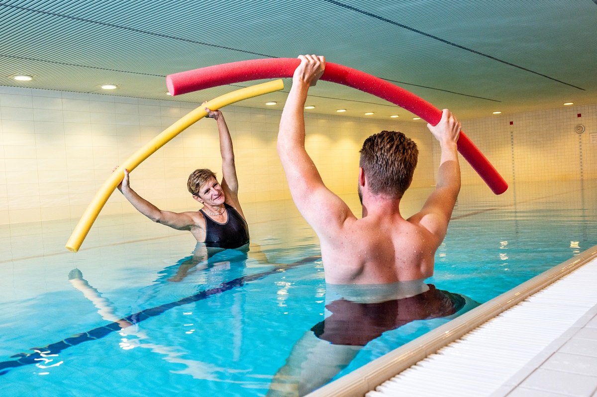 Eine weibliche Patientin und ein männlicher Patient stehen bis zum Brustkorb im Wasser und führen mit einer Poolnudel in den Händen Beweglichkeitsübungen aus.